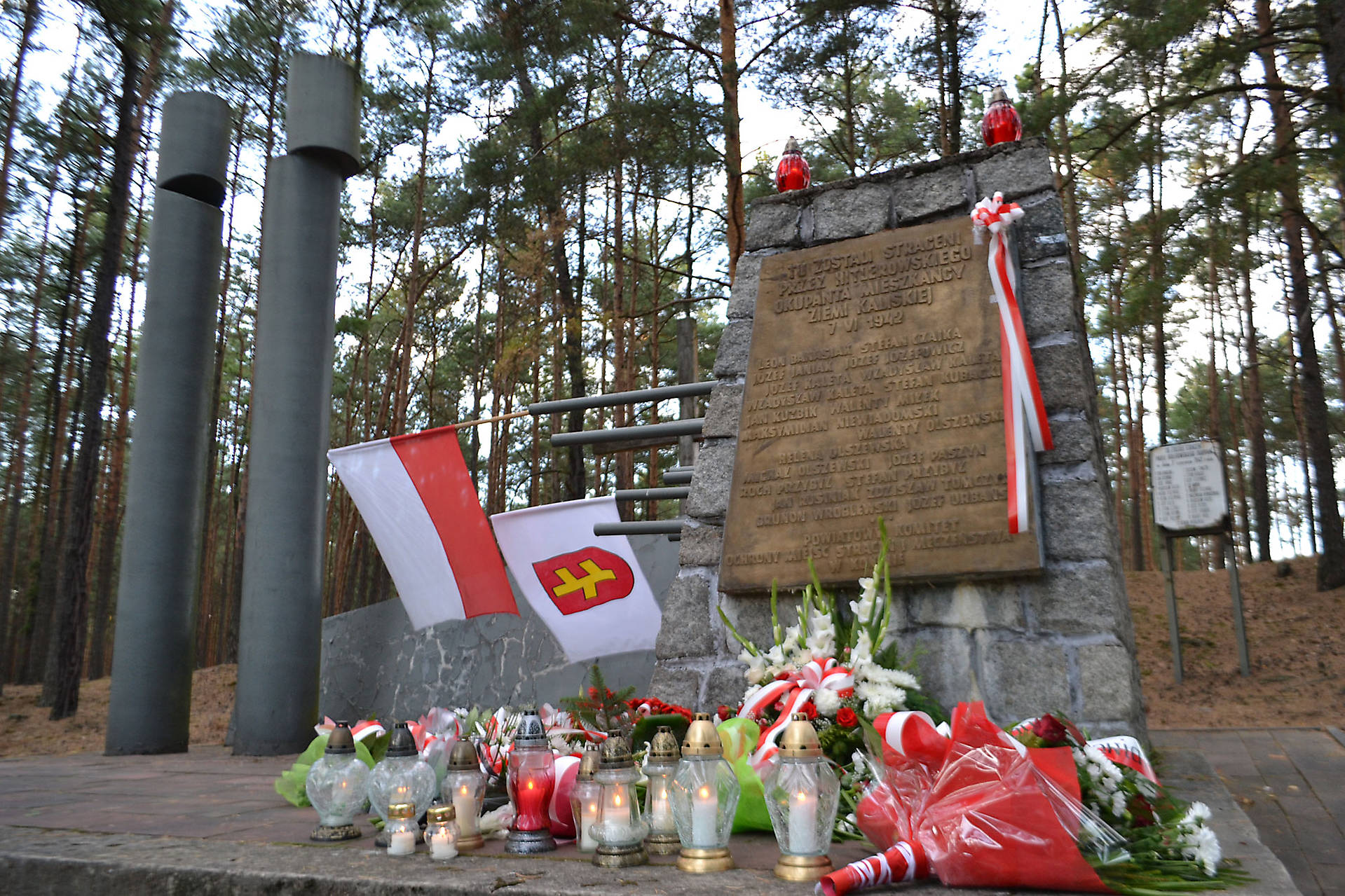 Mogiła w lesie w Białej Panieńskiej_1: Zdjęcie przedstawia udekorowany kwiatami i flagami pomnik wykonany z kamienia.