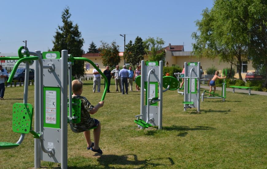Place zabaw_13: Zdjęcie przedstawia dorosłych i dzieci przy urządzeniach do ćwiczeń na tle zieleni