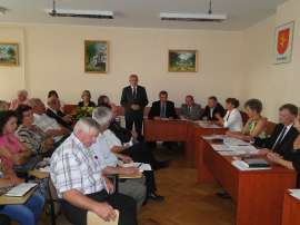 Sesja absolutoryjna_16: Zdjęcie przedstawia siedzących radnych, stojącego burmistrza i obrazy na ścianie.